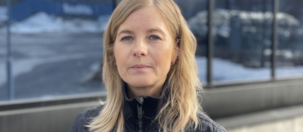 En kvinna med ljust hår som står framför en vägg med massa fönster.