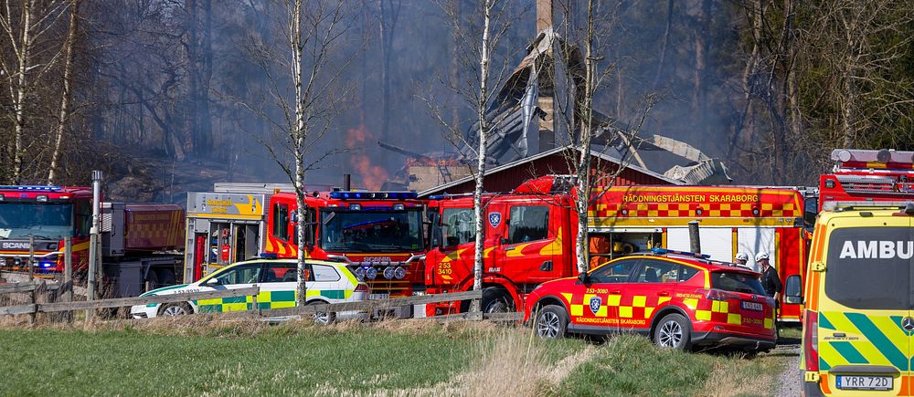 Brandkår och ambulans vid ett husbrand på landsbygden. Röken stiger från det brinnande huset.
