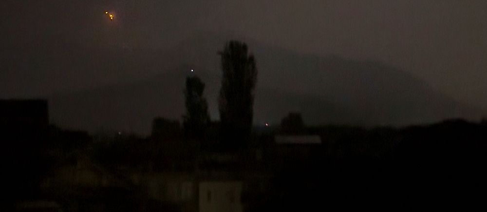 Nattvy över stad med svagt lysande fönster och ett ljussken på himlen i bakgrunden.