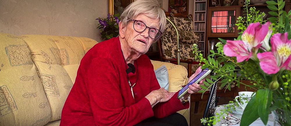 87-åriga Kirsten sitter i en soffa i sitt hem och håller i en mobiltelefon.
