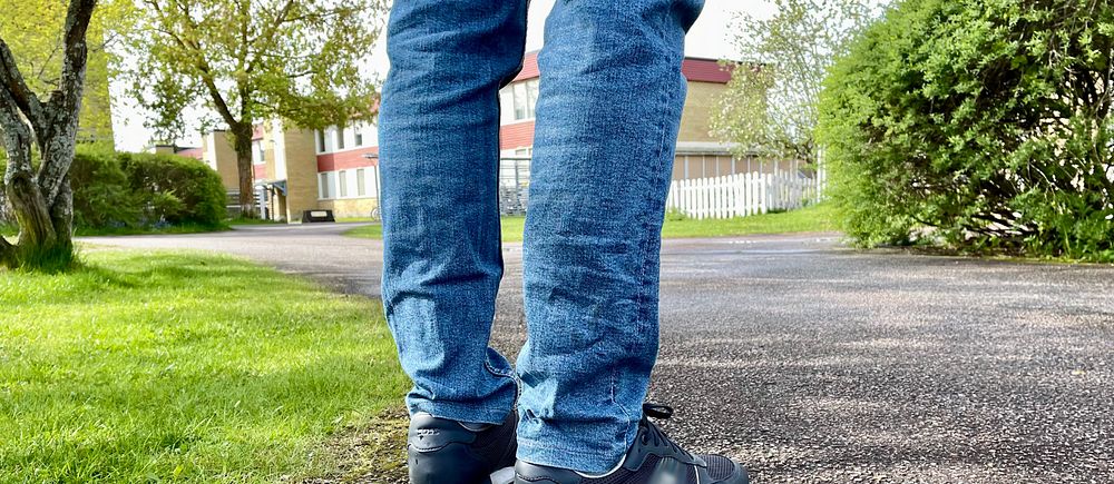 Person i jeans och sneakers står på en asfalterad gångväg i grönskande parkmiljö.
