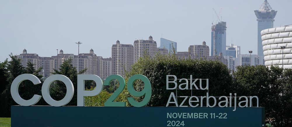 COP29-skylt i Baku, Azerbaijan, med datum 11-22 november 2024, omgiven av grönska och en stadsprofil i bakgrunden.