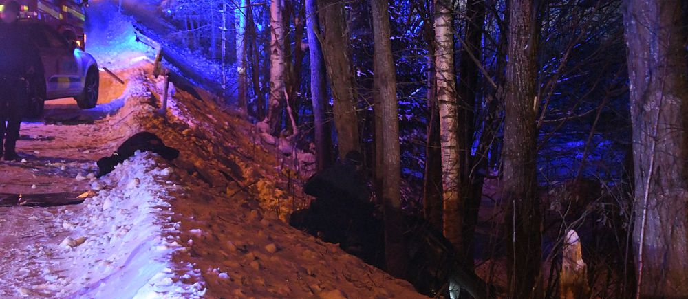 blåljus på snöig väg, en person arbetar vid en bil som ligger i det branta diket bland träd