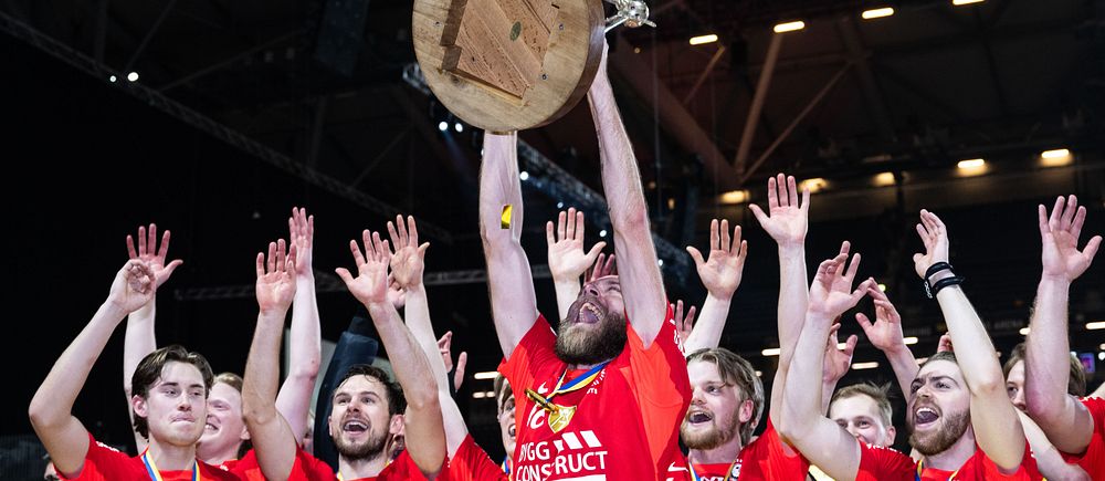 Handbollslag firar med trofé i luften efter vinst. Spelare i röda tröjor jublar.