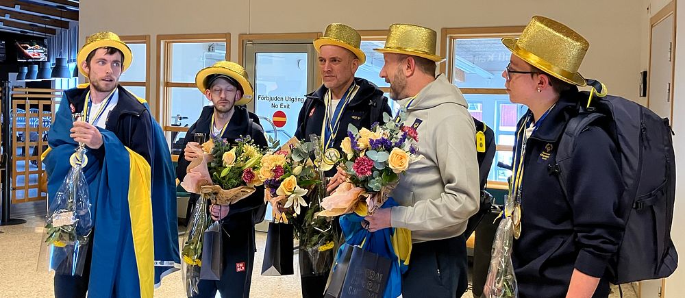 Fem personer med guldhattar står i grupp och håller blommor.