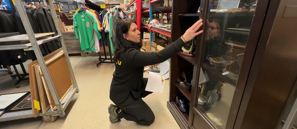 Kvinna i lokal organiserar hyllor med glasdörrar, omgiven av kläder och varor från auktionen av Svennis kvarlåtenskap.