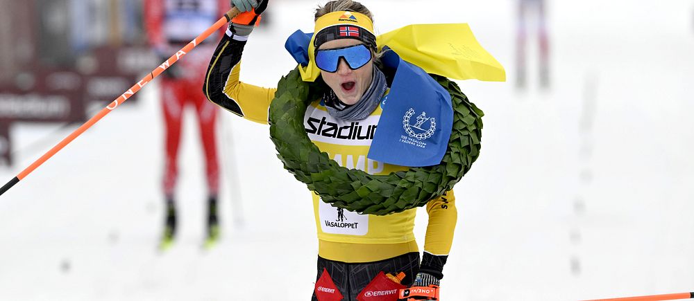 En skidåkare i en gul tävlingsdräkt korsar mållinjen med en laurbärkrans och en gul och blå flagga runt halsen. I bakgrunden syns flera andra skidåkare på en snöig bana.