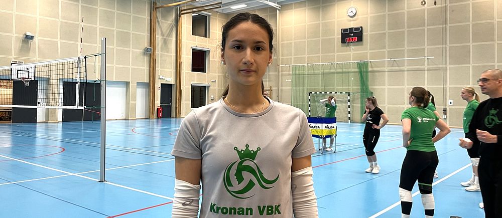 En ung kvinnlig volleybollspelare från Kronan VBK står i borstahusens idrottshall. Tränare och andra volleybollspelare syns i bakgrunden.