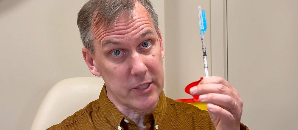 Vetenskapsreporter Johan Bergendorff håller en spruta med blått lock i en klinisk miljö.
