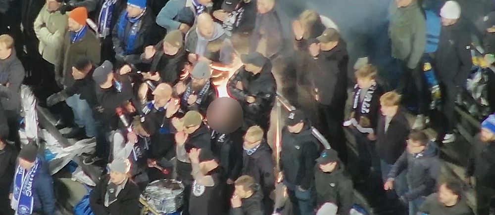 Folksamling klädd i mörka kläder, med någon hållande en fackla, omgiven av rökmoln.