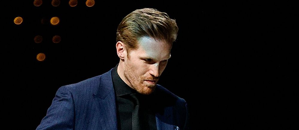 En man i kostym håller en ishockeyklubba mot svart bakgrund.