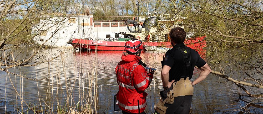 Två räddningsarbetare står vid vattnet och ser mot en röd räddningsbåt.