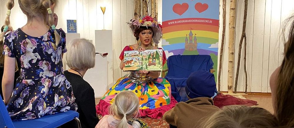 Person klädd i färggrann klänning läser bok för barn på bibliotek. Barnen sitter på golvet.