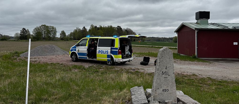 Polisbil med öppna dörrar på landsväg nära röd byggnad och ett milsten. Molnig himmel.