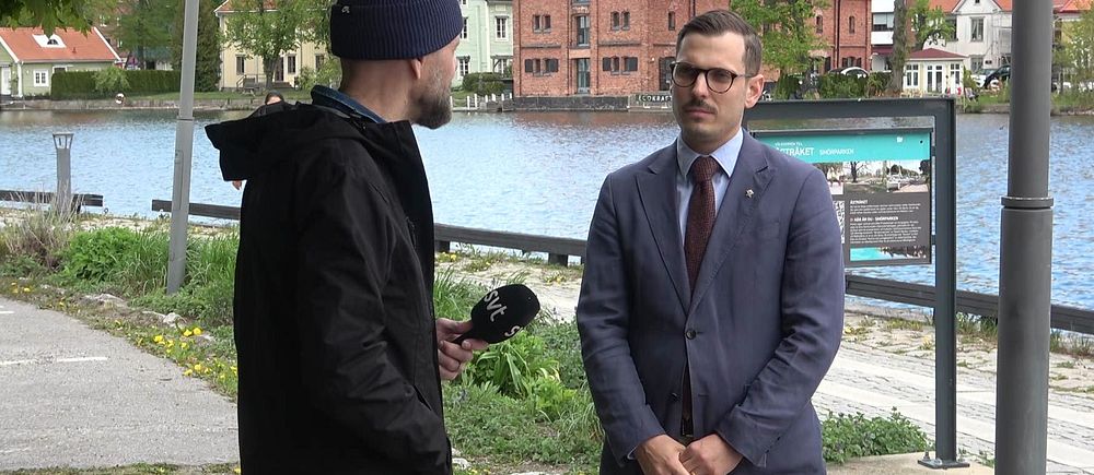 Reporter intervjuar man i kostym vid vattnet med byggnader i bakgrunden.