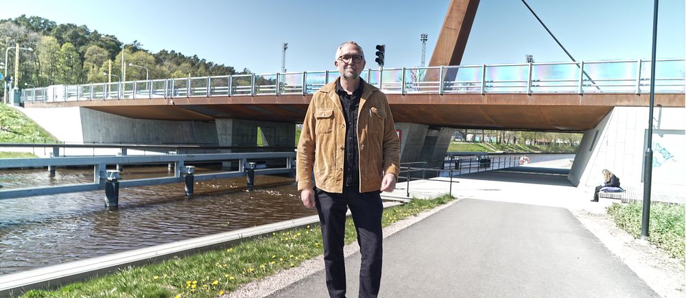 Man i brun jacka står på gångväg framför träbro vid kanal en solig dag.