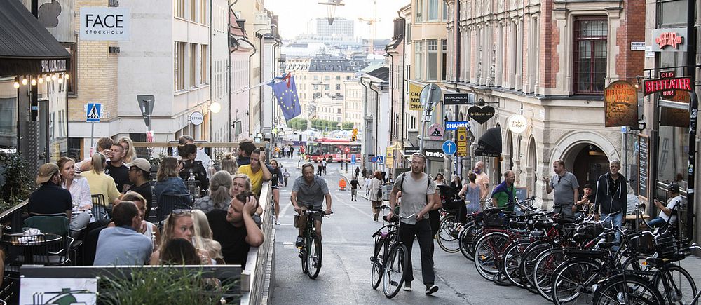 Folk som går och cyklar och fullsatt uteservering i sommarväder på en gata i Stockholm.