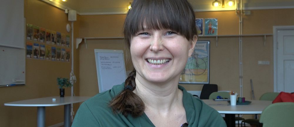 Maria Östlund i grön tröja sitter i ett klassrum. Whiteboard och affischer syns i bakgrunden.