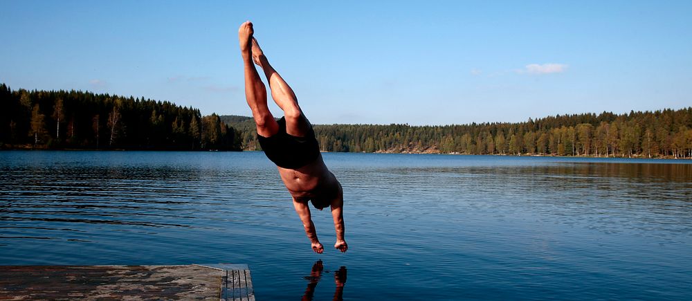 Person dyker från brygga ner i en sjö en solig dag med skog i bakgrunden.