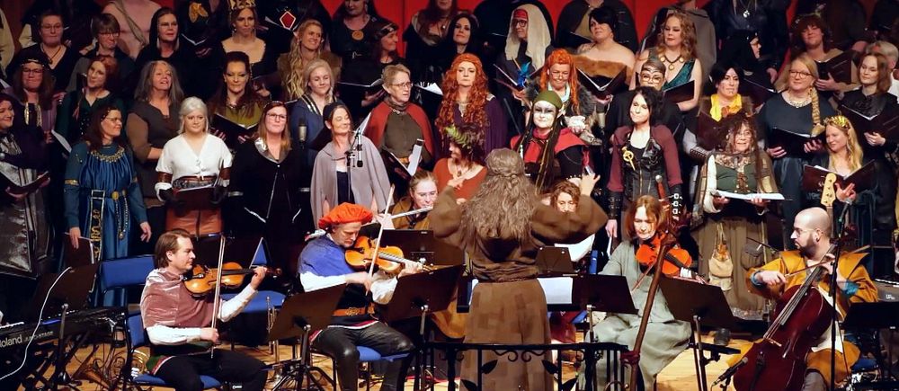 Kör och orkester framför musik tillsammans på scen, många bär fantasifulla kostymer.