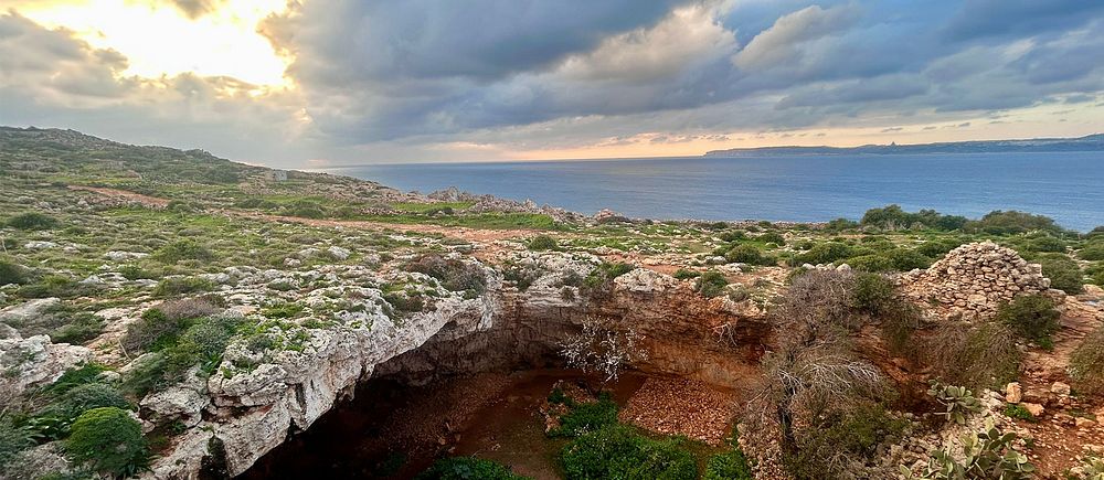 Utsikt över kustlandskap på Malta  med grotta, vegetation och hav under molnigt himmel.