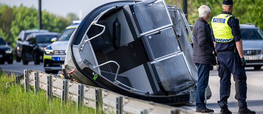 Bil har vält vid vägkant, polis och civilperson står bredvid, kö med bilar i bakgrunden.