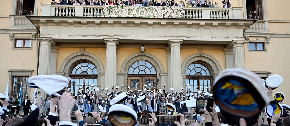 Studenter firar utanför skolbyggnad, viftar med mössor mot balkong.