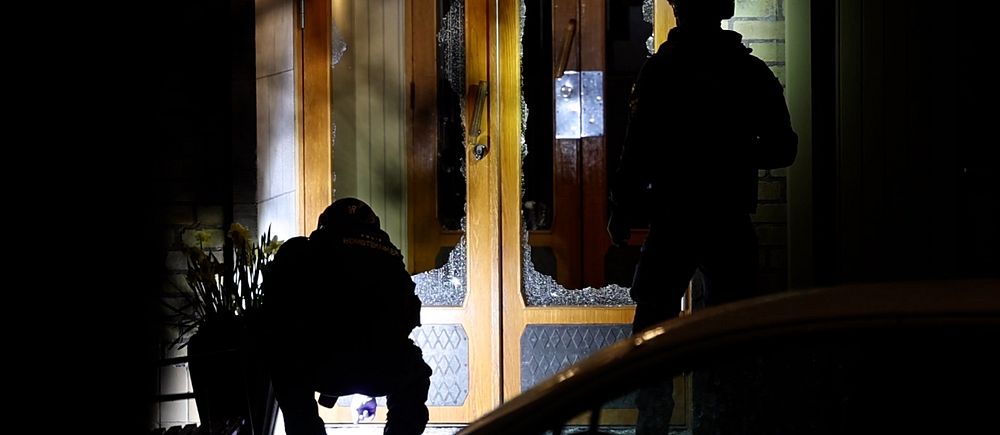 Två poliser i skugga vid en dörr under kvällen, en hukar och en står upprätt.