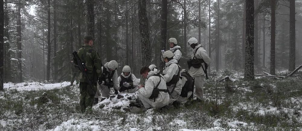 Soldater i vinterkamouflage i skog, diskuterar karta och strategi.