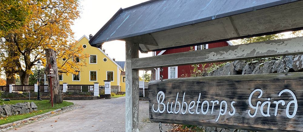 Skylt på Bubbetorps gård med vit text, i bakgrunden ett rött och ett gult hus samt ett höstfärgat träd.