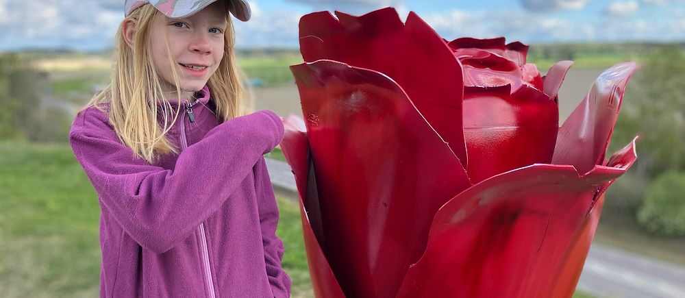 Flicka i tioårsåldern i lila jacka står bredvid en stor röd rosstaty utomhus.