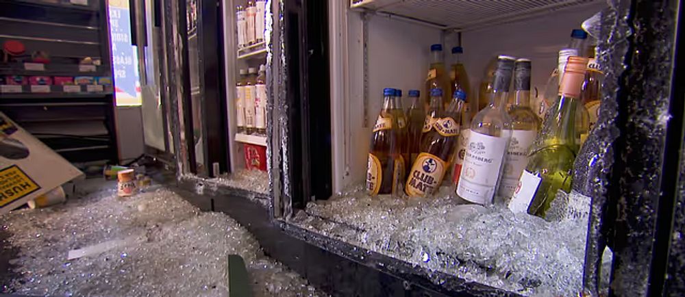 Flaskor bland krossat glas framför trasigt kylskåp i butik.