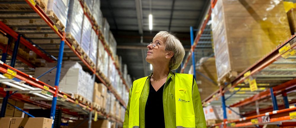 Person i gul väst står i ett lager mellan höga hyllor fyllda med kartonger och paket.