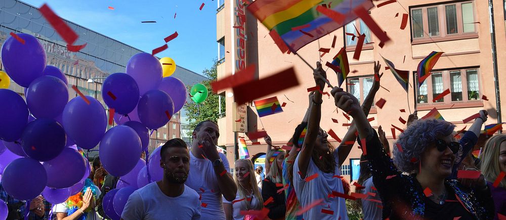 Personer firar Pride med regnbågsflaggor och lila ballonger. Confetti i luften.