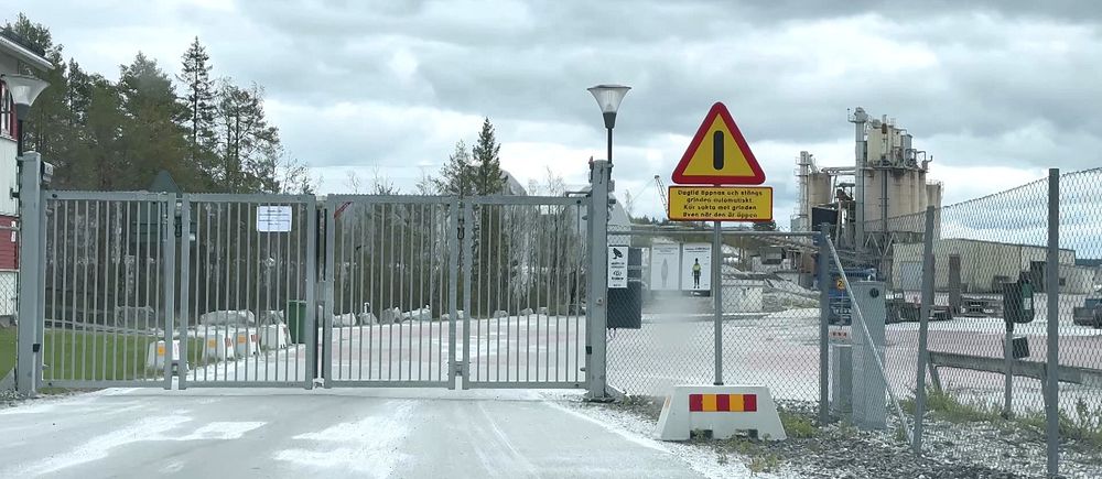 Grind med varningsskylt vid infart till industriområde under molnig himmel.