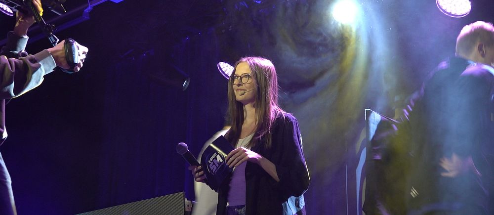 Kvinna talar på scen med stark scenbelysning.