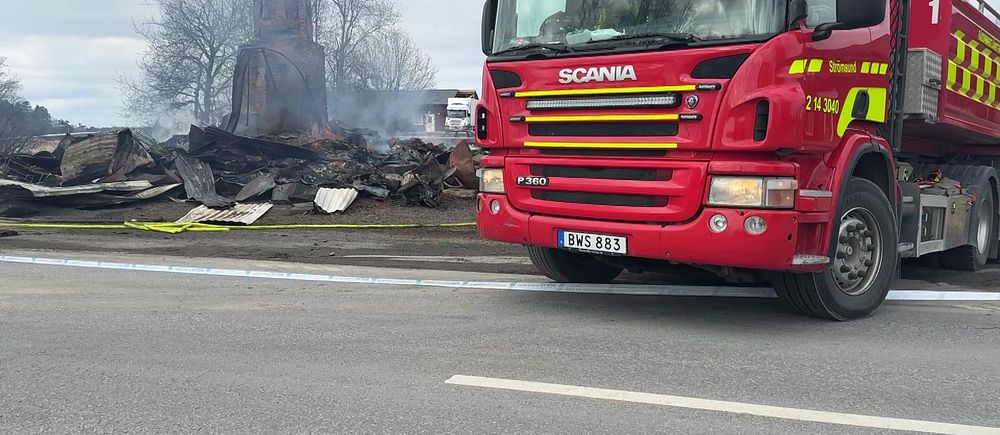 Brandbil framför ruiner efter en brand, rök syns från resterna i bakgrunden.