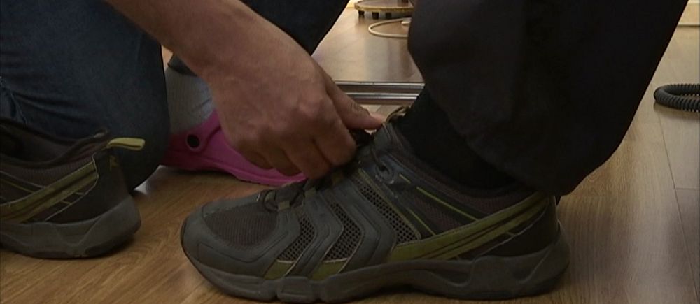 En person knyter skosnören på ett par mörka sneakers inomhus.