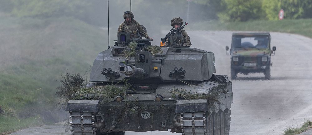 Två soldater sitter på en stridsvagn som kör på landsväg, följd av ett militärfordon.