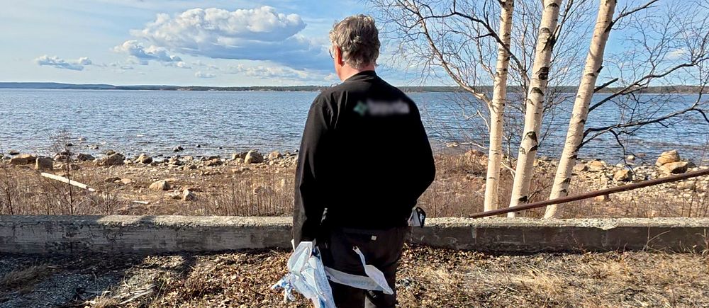 En person med gula stövlar vid en strandkant med träd och vatten i bakgrunden.
