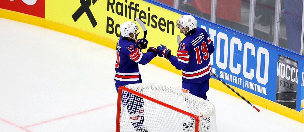 Två hockeyspelare firar mål bakom målbur, målvakt sitter på isen framför dem.