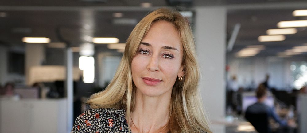 Kvinna med blont hår står på en kontorsarbetsplats med skrivbord och datorer i bakgrunden.