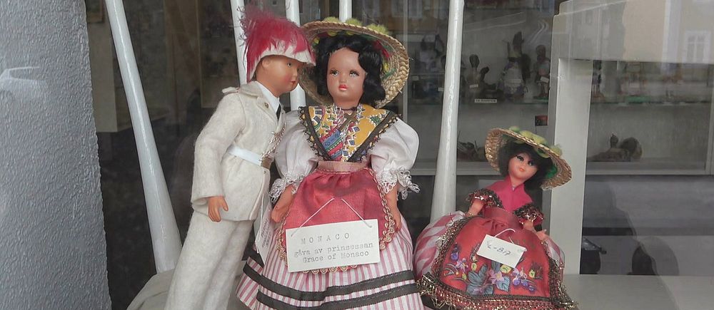 Tre dockor klädda i bationaldräkt uppställda i en monter. På ena dockan finns en skylt att den skänkts av Grace om Monaco