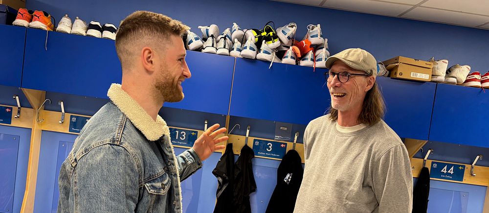 Två personer samtalar i ett omklädningsrum med blåa skåp och sportskor på hyllan.