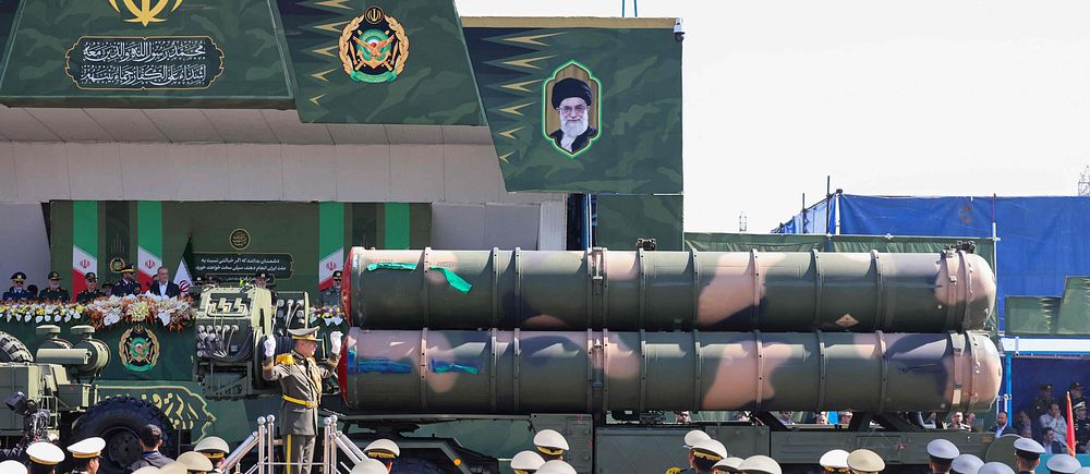 Militärparad i Iran, med soldater och lastbil som bär missiler framför byggnad.