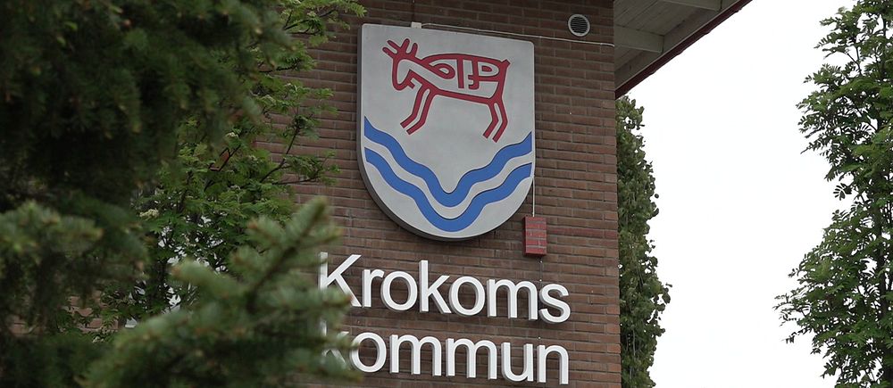 Bild med träd i förgrunden med Krokoms kommunhus i bakgrunden, visar kommunskölden