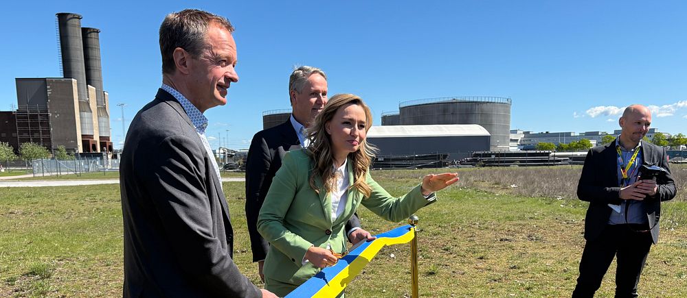 Ebba Busch (KD) i grön kostym klipper blågul band utomhus vid industriområde, tre män närvarande.