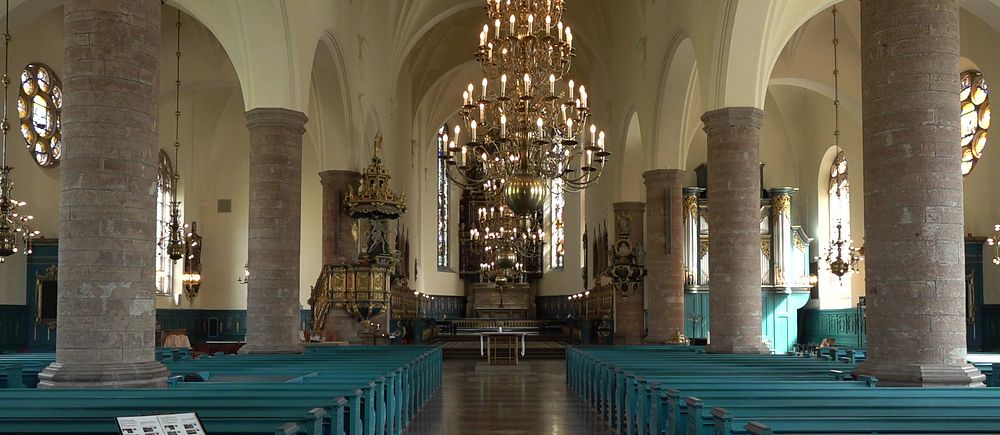 Interiör av kyrka med blå bänkar, höga pelare och stora ljuskronor i taket.