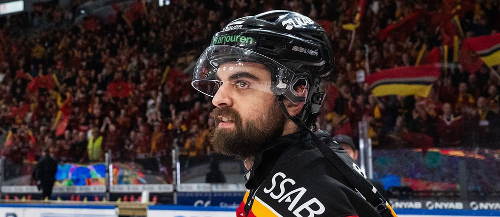 Ishockeyspelare i röd tröja på isen inför en fylld läktare med supportrar och flaggor.