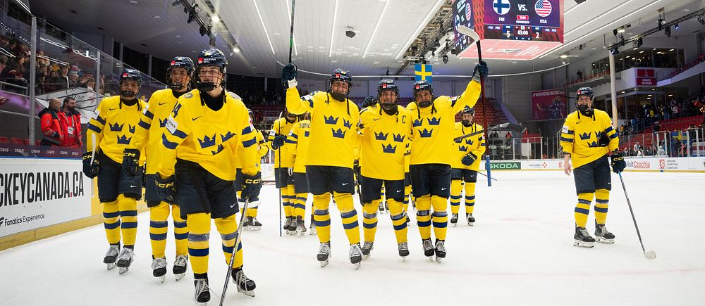 Svenska hockeylaget på isen, klädda i gula tröjor, lyfter sina klubbor i triumf.
